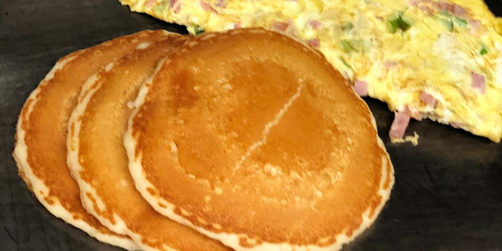 A stack of golden hot cakes on the griddle next to a ham omelet